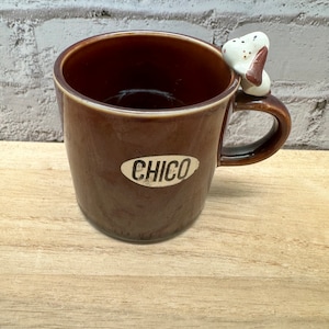 May include: A brown ceramic mug with a small dog figurine attached to the handle. The mug has the word "CHICO" printed on the side.