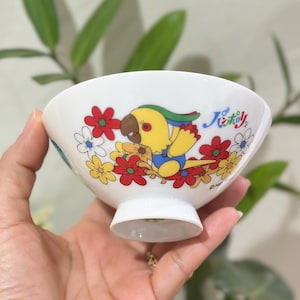 Pu&ograve; includere: Ciotola in ceramica bianca con un'illustrazione colorata di un pappagallo giallo circondato da fiori rossi, gialli e bianchi. La ciotola ha una piccola base a piede e testo giapponese.