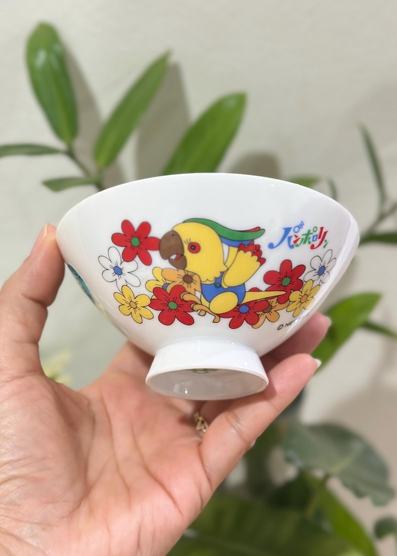 Pu&ograve; includere: Ciotola in ceramica bianca con un'illustrazione colorata di un pappagallo giallo circondato da fiori rossi, gialli e bianchi. La ciotola ha una piccola base a piede e testo giapponese.