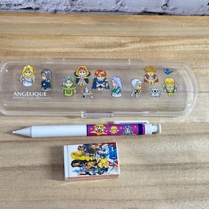 May include: A clear plastic pencil case with cartoon character graphics, a white pen with a purple and pink design, and a small eraser with a colorful illustration. The pencil case has the word "ANGELIQUE" printed on it.