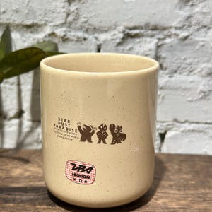 May include: A cream-colored ceramic mug with brown speckles featuring a graphic of three figures and the text STAR DUST PARADISE SINCE 1966 LIVE ACTION PROTECTOR OF THE WORLD SCIENCE FICTION. Below this is a red and black NICHION logo emblem.