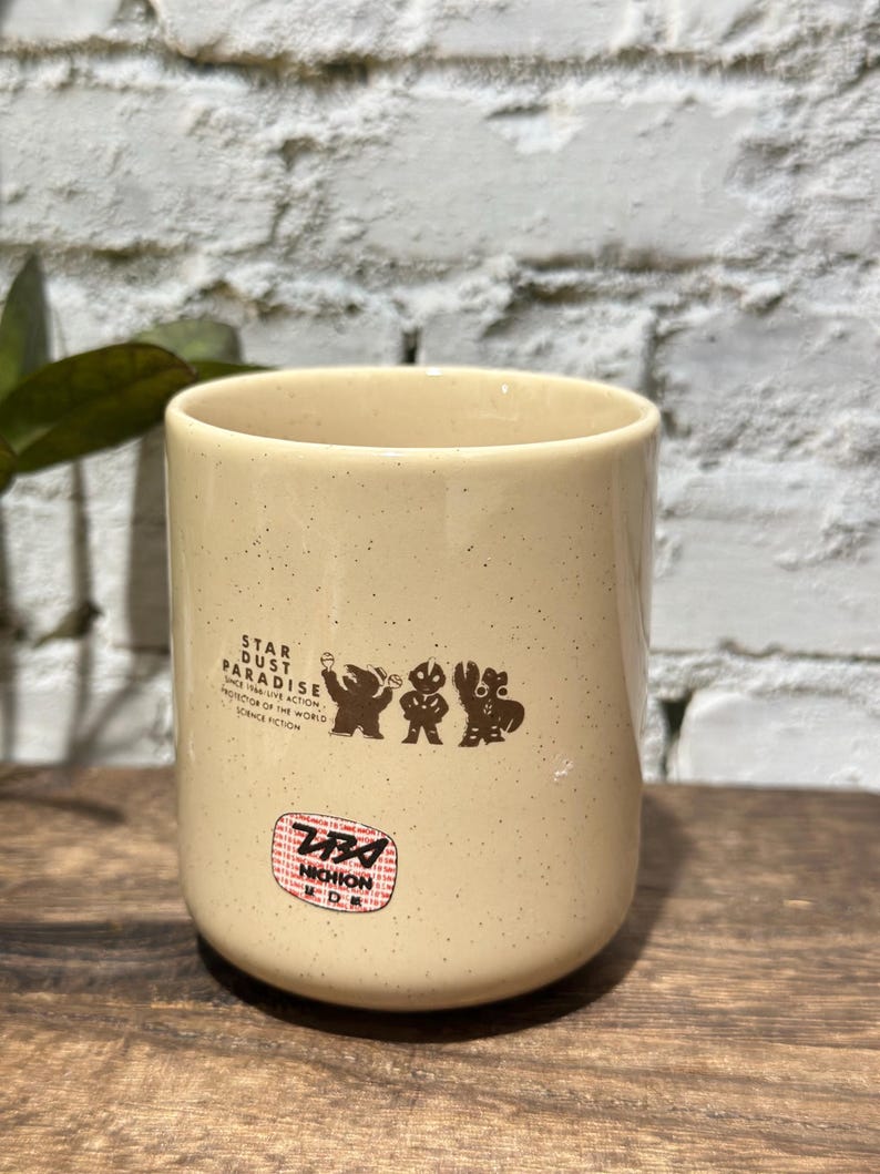 May include: A cream-colored ceramic mug with brown speckles featuring a graphic of three figures and the text STAR DUST PARADISE SINCE 1966 LIVE ACTION PROTECTOR OF THE WORLD SCIENCE FICTION. Below this is a red and black NICHION logo emblem.