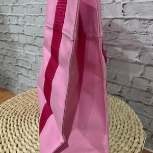 K&ouml;nnte beinhalten: Eine rosa Canvas-Tragetasche mit rosa und roten Henkeln. Die Tasche liegt auf einer geflochtenen Weidenoberfl&auml;che.