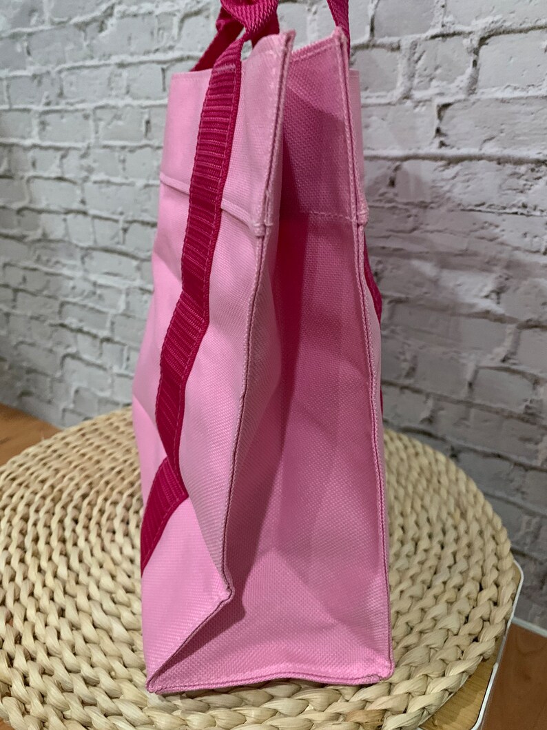 K&ouml;nnte beinhalten: Eine rosa Canvas-Tragetasche mit rosa und roten Henkeln. Die Tasche liegt auf einer geflochtenen Weidenoberfl&auml;che.