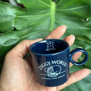 Könnte beinhalten: Eine dunkelblaue Keramiktasse mit einer weißen Comicfigur und dem Text "Ziggy World" und "Universal Press Syndicate".