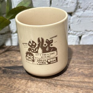 May include: A cream-colored ceramic mug with brown speckled details and a graphic print featuring cartoon characters. The text reads: HOPPING! Hop Step and Jump, POPPING! Nuts, LET'S SING A SONG WITH ME!, JOLLY TIME, and UTM SONG.
