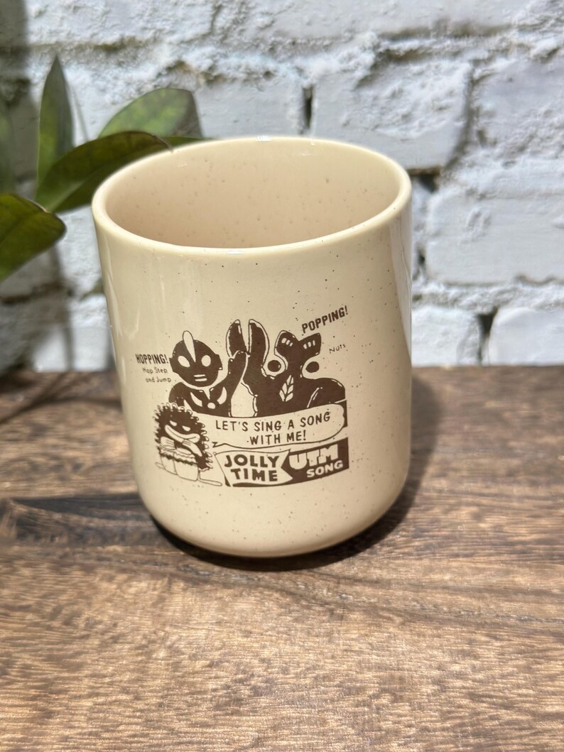May include: A cream-colored ceramic mug with brown speckled details and a graphic print featuring cartoon characters. The text reads: HOPPING! Hop Step and Jump, POPPING! Nuts, LET'S SING A SONG WITH ME!, JOLLY TIME, and UTM SONG.