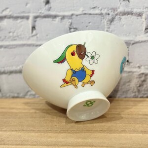 Pu&ograve; includere: Ciotola in ceramica bianca con un disegno di pappagallo dei cartoni animati. Il pappagallo &egrave; giallo con un becco marrone e indossa una tuta blu. Tiene in mano un fiore bianco. La ciotola ha una base a piede e un disegno azzurro chiaro sul lato.