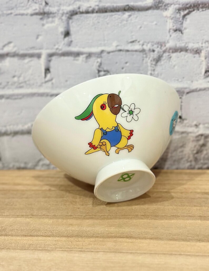 Pu&ograve; includere: Ciotola in ceramica bianca con un disegno di pappagallo dei cartoni animati. Il pappagallo &egrave; giallo con un becco marrone e indossa una tuta blu. Tiene in mano un fiore bianco. La ciotola ha una base a piede e un disegno azzurro chiaro sul lato.