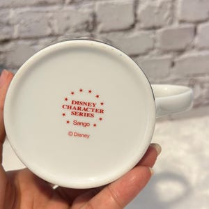 Puede incluir: Taza de cer&aacute;mica blanca con asa, con texto rojo que dice "DISNEY CHARACTER SERIES" y "Sango". El logotipo de Disney tambi&eacute;n es visible. La taza est&aacute; sujeta por una mano, con un fondo de ladrillos blancos.