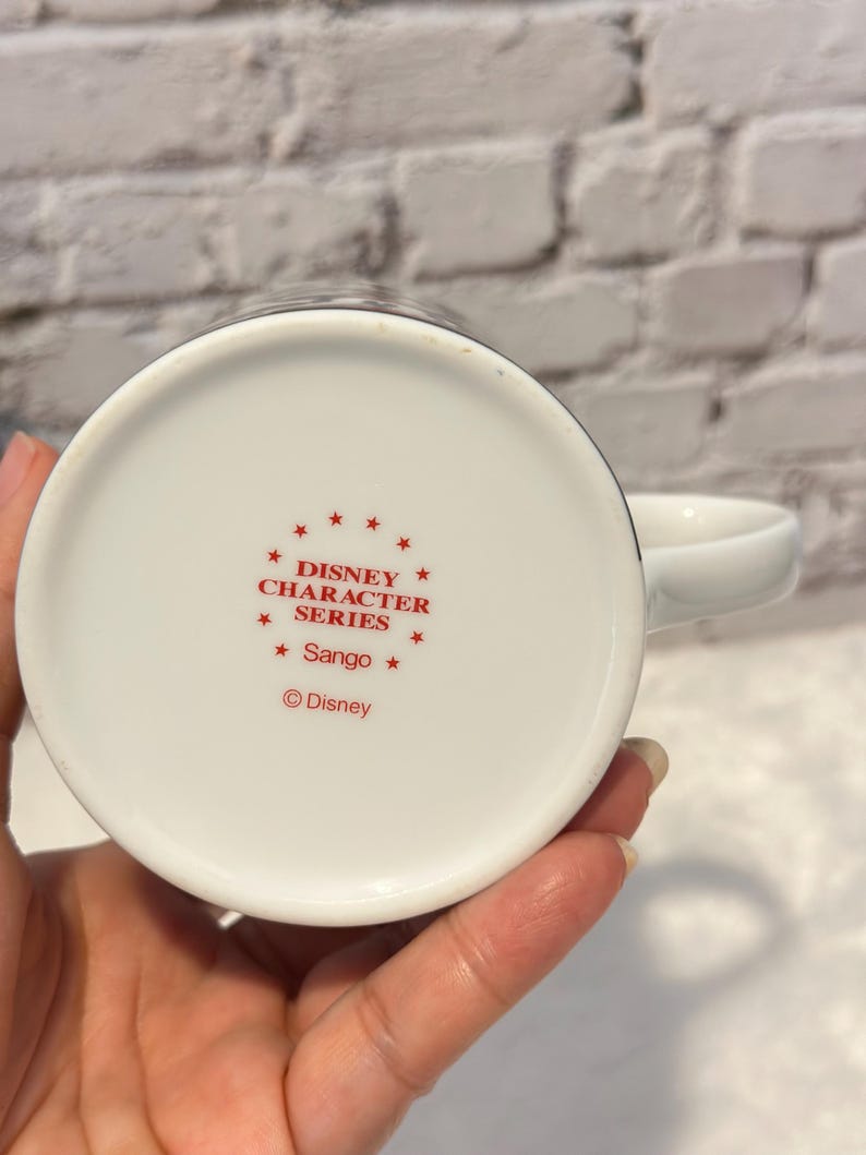 Puede incluir: Taza de cer&aacute;mica blanca con asa, con texto rojo que dice "DISNEY CHARACTER SERIES" y "Sango". El logotipo de Disney tambi&eacute;n es visible. La taza est&aacute; sujeta por una mano, con un fondo de ladrillos blancos.