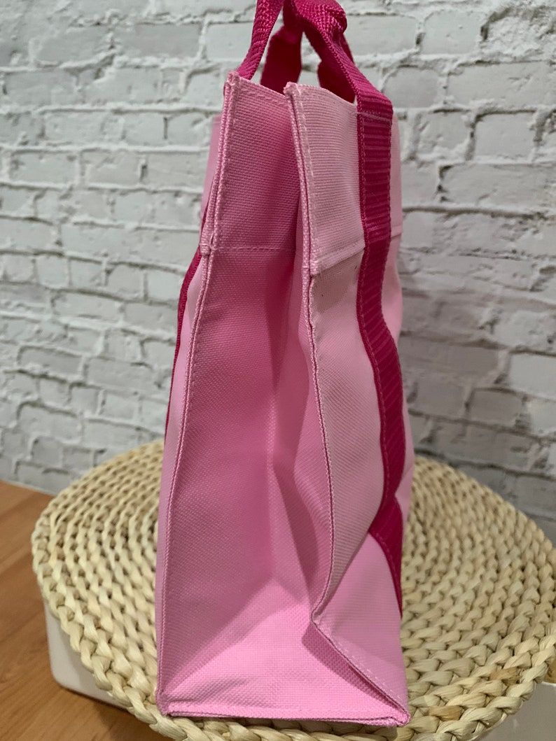 K&ouml;nnte beinhalten: Eine rosa und fuchsiafarbene Tragetasche mit einem einzigen Henkel. Die Tasche ist aus Canvas gefertigt und hat ein schlichtes Design.