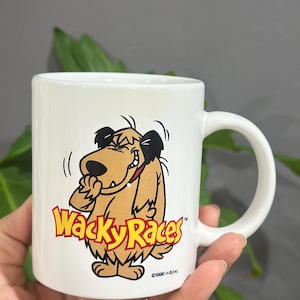 Może przedstawiać: Biały ceramiczny kubek z postacią psa z kreskówki Wacky Races. Pies jest brązowy z czarnymi uszami i czerwoną obrożą, a kubek ma logo programu w kolorze żółtym i czerwonym. Kubek ma standardowy rozmiar z zakrzywionym uchwytem.