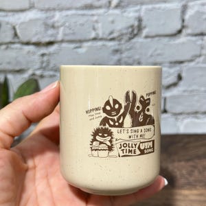 May include: A cream-colored ceramic mug with a speckled finish featuring brown graphic illustrations of Ultraman characters. The text reads: HOPPING! Hop Step and Jump, POPPING!, LET'S SING A SONG WITH ME!, and JOLLY TIME UTH SONG.