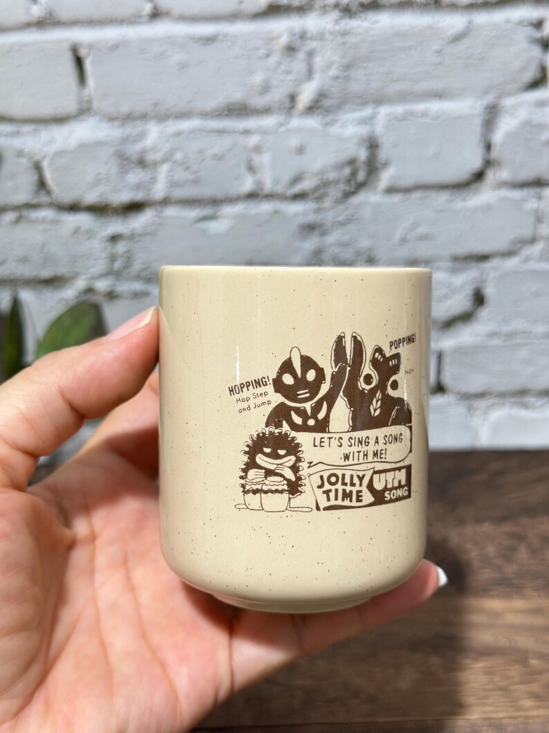 May include: A cream-colored ceramic mug with a speckled finish featuring brown graphic illustrations of Ultraman characters. The text reads: HOPPING! Hop Step and Jump, POPPING!, LET'S SING A SONG WITH ME!, and JOLLY TIME UTH SONG.