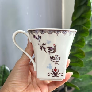 May include: White ceramic mug with a brown and purple floral design and a small red crown. The mug has a handle and a square base. The design includes a bird, hearts, and a Mickey Mouse silhouette.
