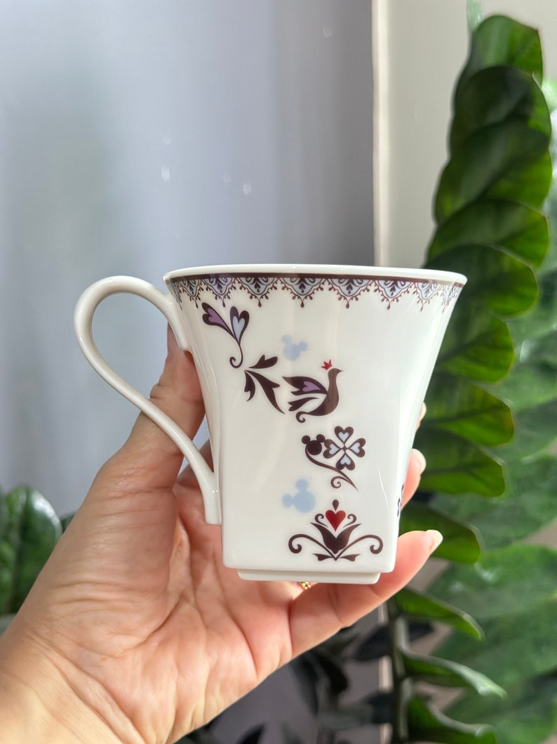 May include: White ceramic mug with a brown and purple floral design and a small red crown. The mug has a handle and a square base. The design includes a bird, hearts, and a Mickey Mouse silhouette.