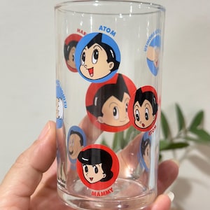 May include: Clear glass tumbler featuring cartoon character Atom and other characters in red and blue circles. The glass is held by a hand, and the design is repeated around the glass. The word "Atom" is visible on the glass.