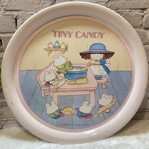 May include: A round, pink metal tray with a cartoon illustration. The illustration features a girl in a blue dress and hat, and several mouse characters. The text "TINY CANDY" is printed at the top of the tray.