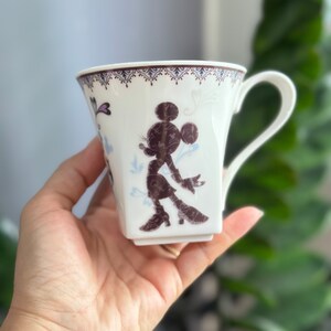May include: White ceramic mug with a brown silhouette of Mickey Mouse on the front. The mug has a decorative border around the top and a handle.