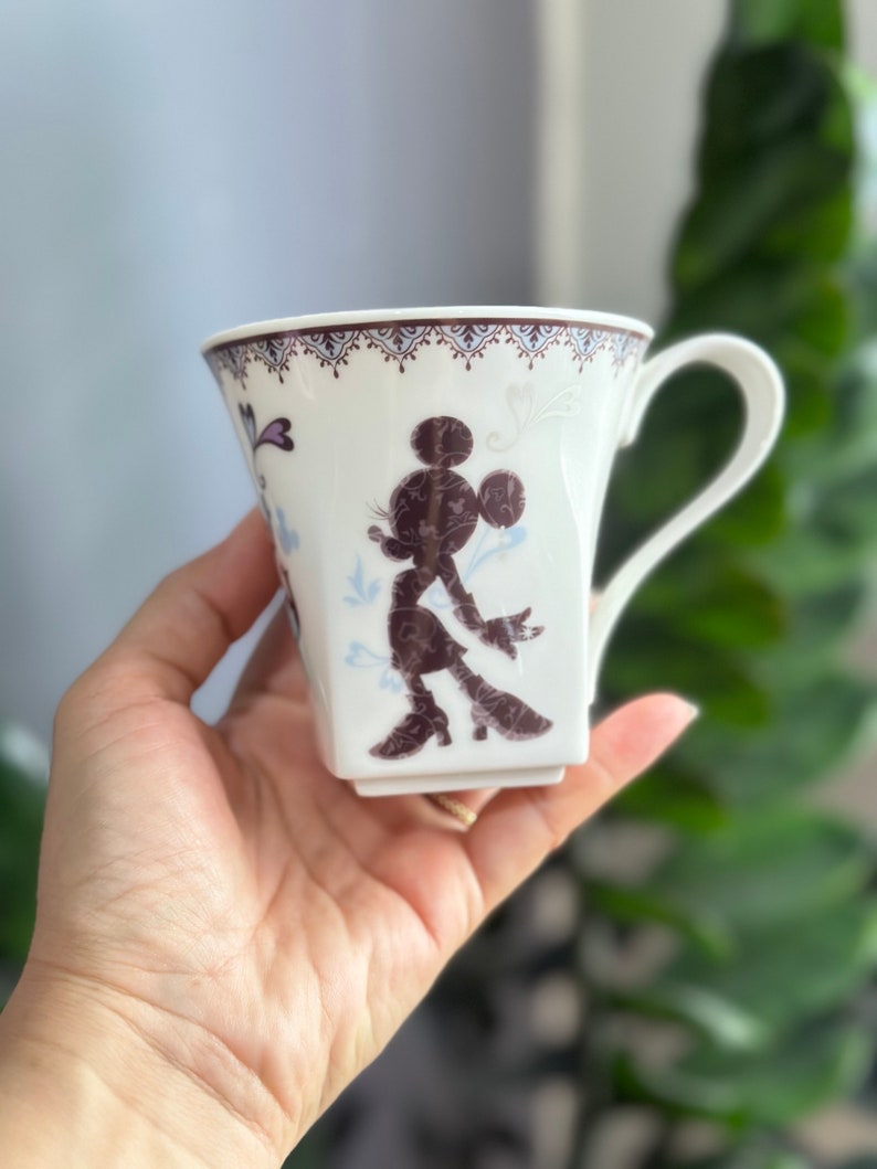 May include: White ceramic mug with a brown silhouette of Mickey Mouse on the front. The mug has a decorative border around the top and a handle.