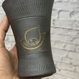 Könnte beinhalten: Eine schwarze Keramiktasse mit einem goldenen Design eines Comic-Tieres in einem Blatt. Der Text "うっしっし" ist in Gold auf dem Boden der Tasse geschrieben.