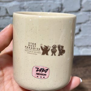 May include: A cream-colored ceramic cup with brown speckles, featuring a brown graphic of three figures and the text STAR DUST PARADISE SINCE 1966 LIVE ACTION PROTECTOR OF THE WORLD SCIENCE FICTION. Below is a red and white circular sticker with NICHION.