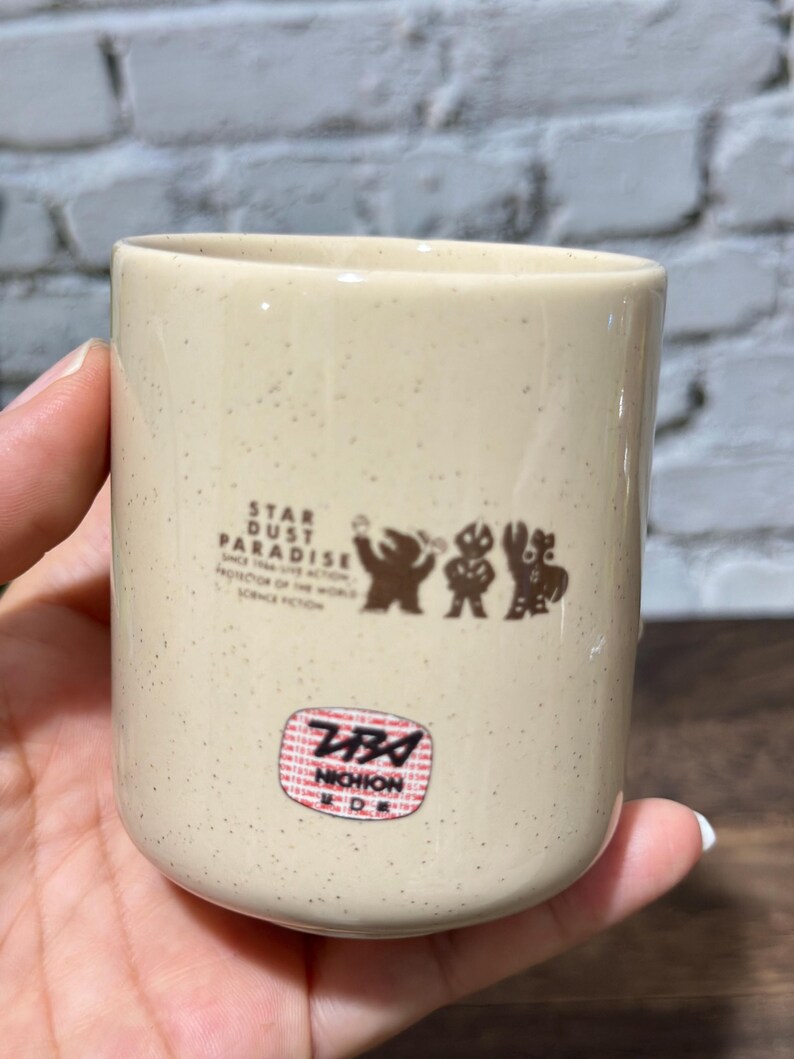 May include: A cream-colored ceramic cup with brown speckles, featuring a brown graphic of three figures and the text STAR DUST PARADISE SINCE 1966 LIVE ACTION PROTECTOR OF THE WORLD SCIENCE FICTION. Below is a red and white circular sticker with NICHION.