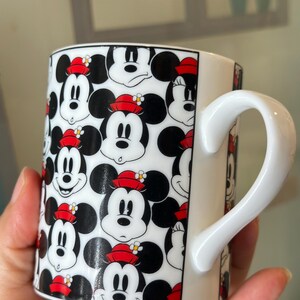 Puede incluir: Taza de cer&aacute;mica blanca con un patr&oacute;n repetido de caras de Mickey Mouse con sombreros rojos y orejas negras. La taza tiene un asa blanca y un borde negro. Un dise&ntilde;o alegre para los fan&aacute;ticos de Disney.
