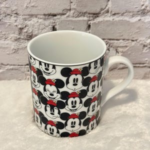 Puede incluir: Taza de cer&aacute;mica blanca con un patr&oacute;n repetido de caras de Mickey Mouse con sombreros rojos. La taza tiene un interior blanco y un asa curva. El dise&ntilde;o incluye colores negro, rojo y blanco, creando un tema l&uacute;dico y reconocible.