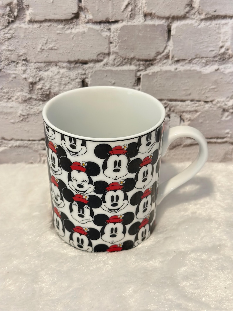 Puede incluir: Taza de cer&aacute;mica blanca con un patr&oacute;n repetido de caras de Mickey Mouse con sombreros rojos. La taza tiene un interior blanco y un asa curva. El dise&ntilde;o incluye colores negro, rojo y blanco, creando un tema l&uacute;dico y reconocible.