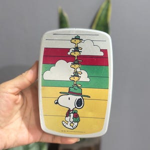 May include: Rectangular, silver-colored lunchbox featuring a Snoopy design. Snoopy is depicted wearing a green hat and backpack, with Woodstock and other birds stacked on top. The background has horizontal stripes of yellow, green, red, and white.