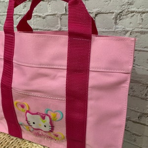 K&ouml;nnte beinhalten: Eine rosa Canvas-Tragetasche mit rosa Henkeln und einem Hello-Kitty-Design mit einem rosa Hello Kitty mit Blumen und Schmetterlingen. Die Tasche hat eine kleine Tasche auf der Vorderseite.