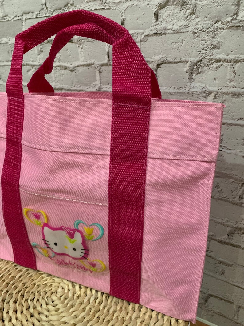 K&ouml;nnte beinhalten: Eine rosa Canvas-Tragetasche mit rosa Henkeln und einem Hello-Kitty-Design mit einem rosa Hello Kitty mit Blumen und Schmetterlingen. Die Tasche hat eine kleine Tasche auf der Vorderseite.