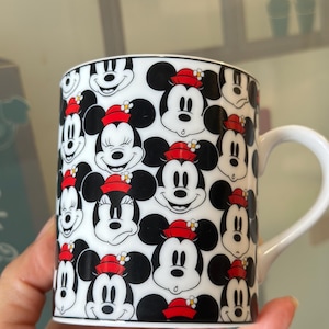 Puede incluir: Taza de cer&aacute;mica blanca con un patr&oacute;n repetido de caras de Mickey Mouse y Minnie Mouse. Los personajes son en blanco, negro y rojo, con Minnie usando un sombrero rojo con una flor. La taza tiene un asa blanca.