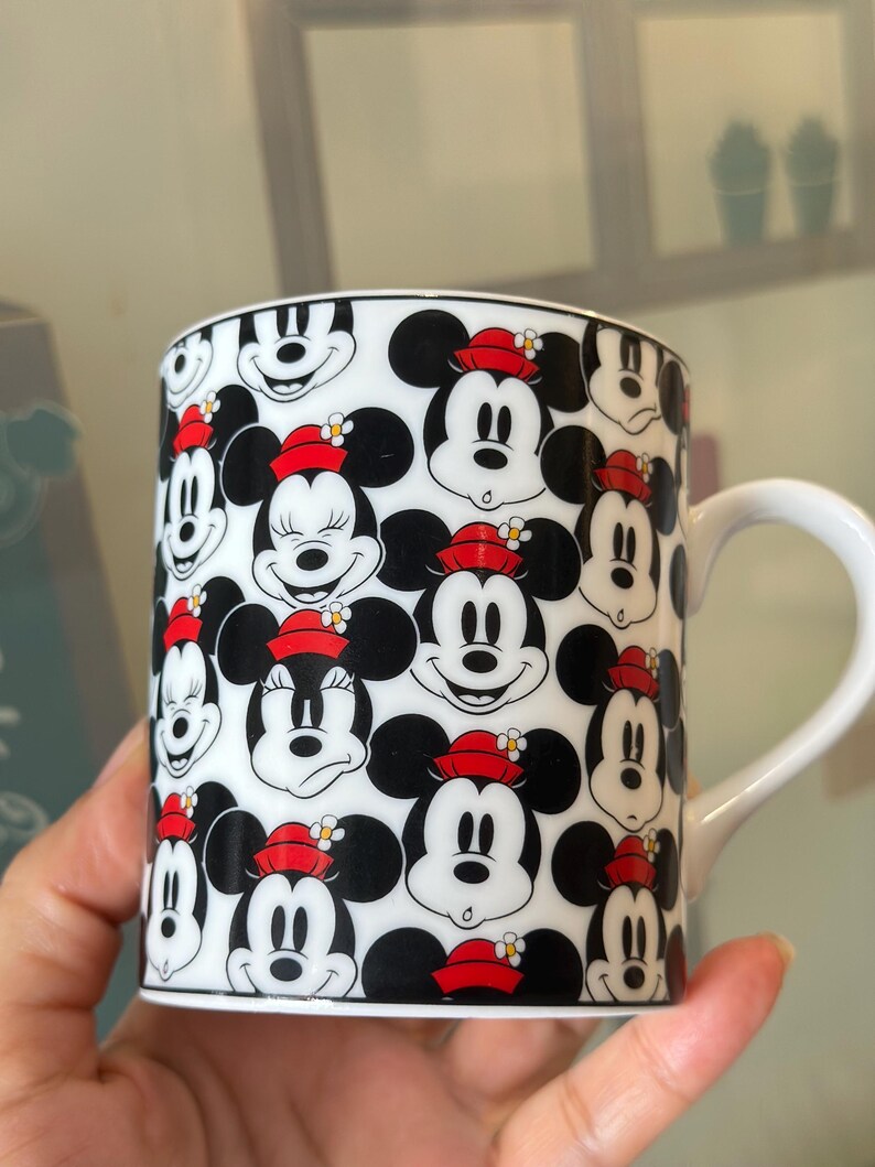 Puede incluir: Taza de cer&aacute;mica blanca con un patr&oacute;n repetido de caras de Mickey Mouse y Minnie Mouse. Los personajes son en blanco, negro y rojo, con Minnie usando un sombrero rojo con una flor. La taza tiene un asa blanca.