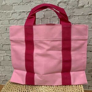 K&ouml;nnte beinhalten: Eine rosa Canvas-Tragetasche mit zwei dicken rosa Henkeln. Die Tasche hat eine einzelne Tasche auf der Vorderseite.