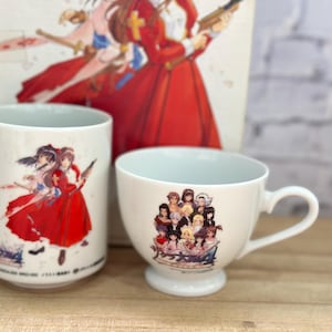 May include: Two white ceramic mugs with anime-style illustrations. One is a tall cylindrical mug, the other a teacup with a handle. The illustrations feature characters in red and white clothing. A box with a similar design is in the background.