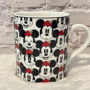 Puede incluir: Taza de cer&aacute;mica blanca con un patr&oacute;n repetido de caras de Mickey Mouse y Minnie Mouse. Los personajes tienen orejas negras, caras sonrientes y llevan sombreros rojos. La taza tiene un asa blanca y un borde negro.