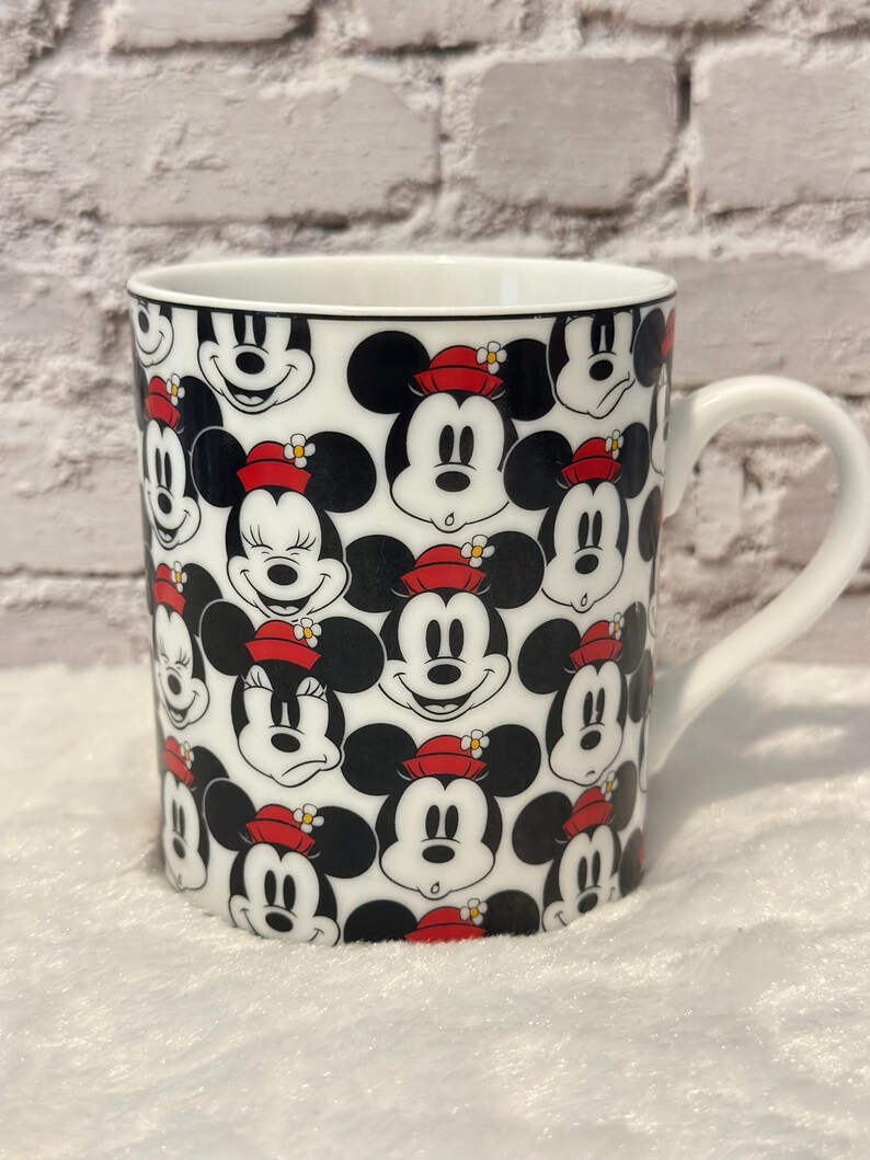Puede incluir: Taza de cer&aacute;mica blanca con un patr&oacute;n repetido de caras de Mickey Mouse y Minnie Mouse. Los personajes tienen orejas negras, caras sonrientes y llevan sombreros rojos. La taza tiene un asa blanca y un borde negro.