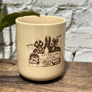 May include: A cream-colored ceramic mug with a speckled finish featuring a brown graphic print of Ultraman characters. The text reads: HOPPING! Hop Step and Jump, POPPING!, LET'S SING A SONG WITH ME! JOLLY TIME UTM SONG.