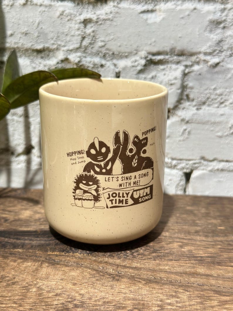 May include: A cream-colored ceramic mug with a speckled finish featuring a brown graphic print of Ultraman characters. The text reads: HOPPING! Hop Step and Jump, POPPING!, LET'S SING A SONG WITH ME! JOLLY TIME UTM SONG.
