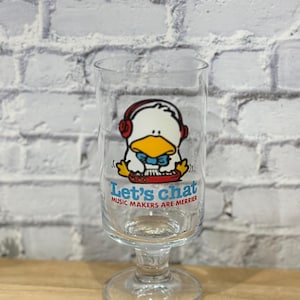 May include: A clear glass with a stem and a white duck wearing headphones and a blue bow tie playing a keyboard. The glass has the text "Let's chat Music Makers Are Merrier" printed on it.