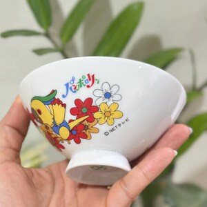 Pu&ograve; includere: Ciotola in ceramica bianca con un personaggio dei cartoni animati e un design floreale. La ciotola presenta un'illustrazione colorata di un personaggio con accenti gialli, blu e verdi, insieme a fiori rossi, gialli e bianchi. &Egrave; visibile anche del testo giapponese.