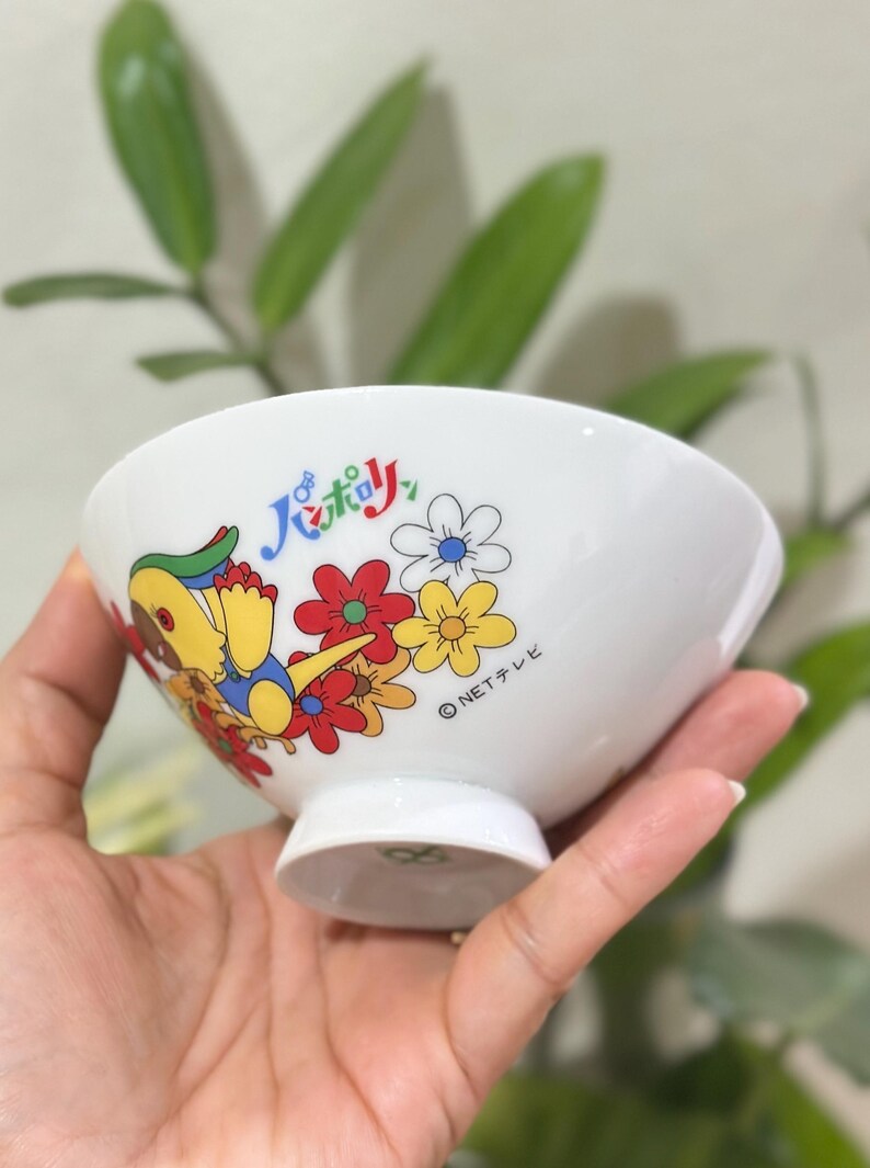 Pu&ograve; includere: Ciotola in ceramica bianca con un personaggio dei cartoni animati e un design floreale. La ciotola presenta un'illustrazione colorata di un personaggio con accenti gialli, blu e verdi, insieme a fiori rossi, gialli e bianchi. &Egrave; visibile anche del testo giapponese.