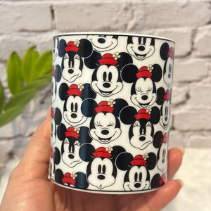 Puede incluir: Una taza de cer&aacute;mica blanca con un patr&oacute;n repetido de caras de Mickey y Minnie Mouse. Los personajes de dibujos animados son en blanco, negro y rojo, con Minnie usando un sombrero rojo con una flor amarilla.