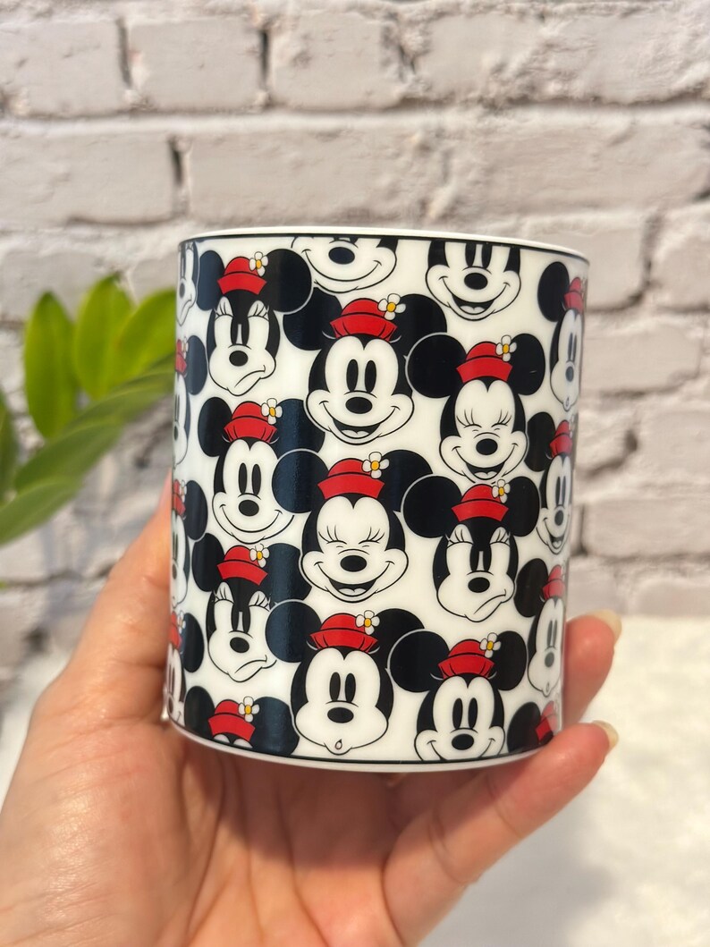 Puede incluir: Una taza de cer&aacute;mica blanca con un patr&oacute;n repetido de caras de Mickey y Minnie Mouse. Los personajes de dibujos animados son en blanco, negro y rojo, con Minnie usando un sombrero rojo con una flor amarilla.