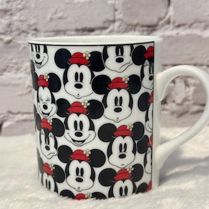 Puede incluir: Taza de cer&aacute;mica blanca con un patr&oacute;n repetido de caras de Mickey Mouse. Las caras tienen orejas negras, sombreros rojos y caras blancas. La taza tiene un asa blanca y un borde negro. Fondo de pared de ladrillo blanco.