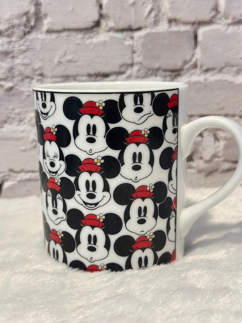 Puede incluir: Taza de cer&aacute;mica blanca con un patr&oacute;n repetido de caras de Mickey Mouse. Las caras tienen orejas negras, sombreros rojos y caras blancas. La taza tiene un asa blanca y un borde negro. Fondo de pared de ladrillo blanco.