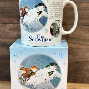May include: A white ceramic mug and matching box featuring illustrations from "The Snowman." The mug depicts a snowman with a boy, and the box shows the same image. The text "The Snowman" is printed on both.
